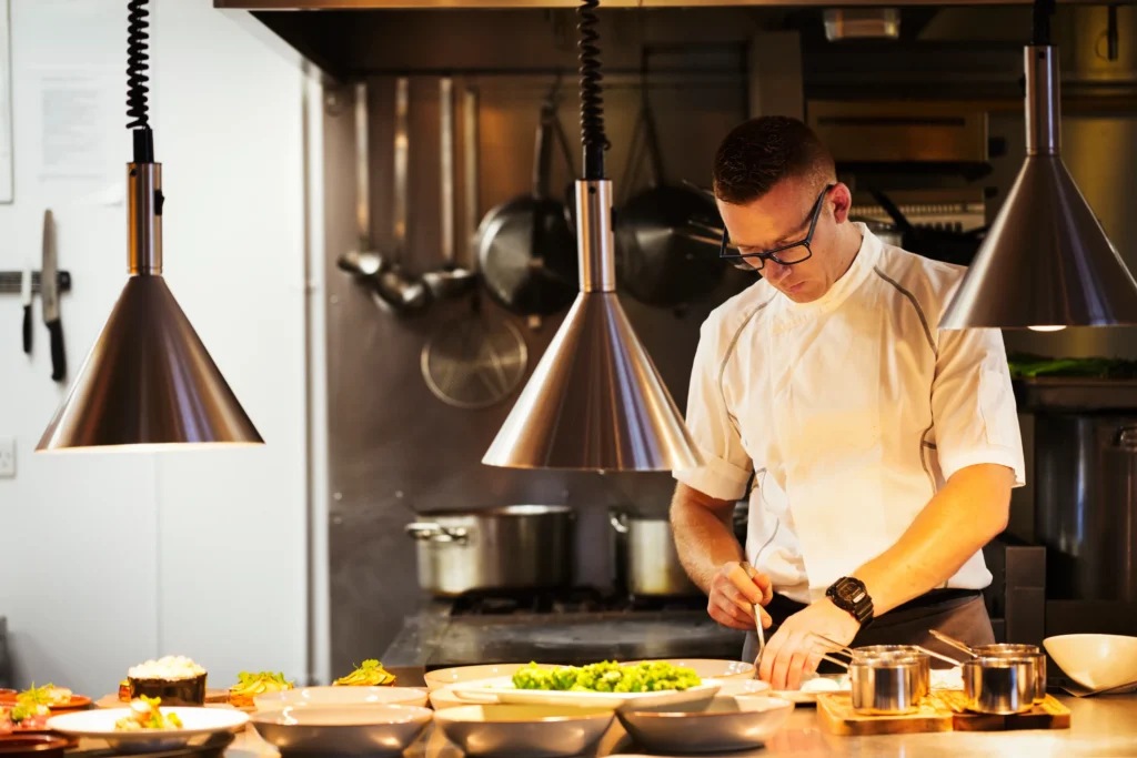 A chef in a white uniform prepares food in a professional kitchen, surrounded by plates of ingredients and metal pendant lights overhead. Fiamma Sun Valley | Italian
