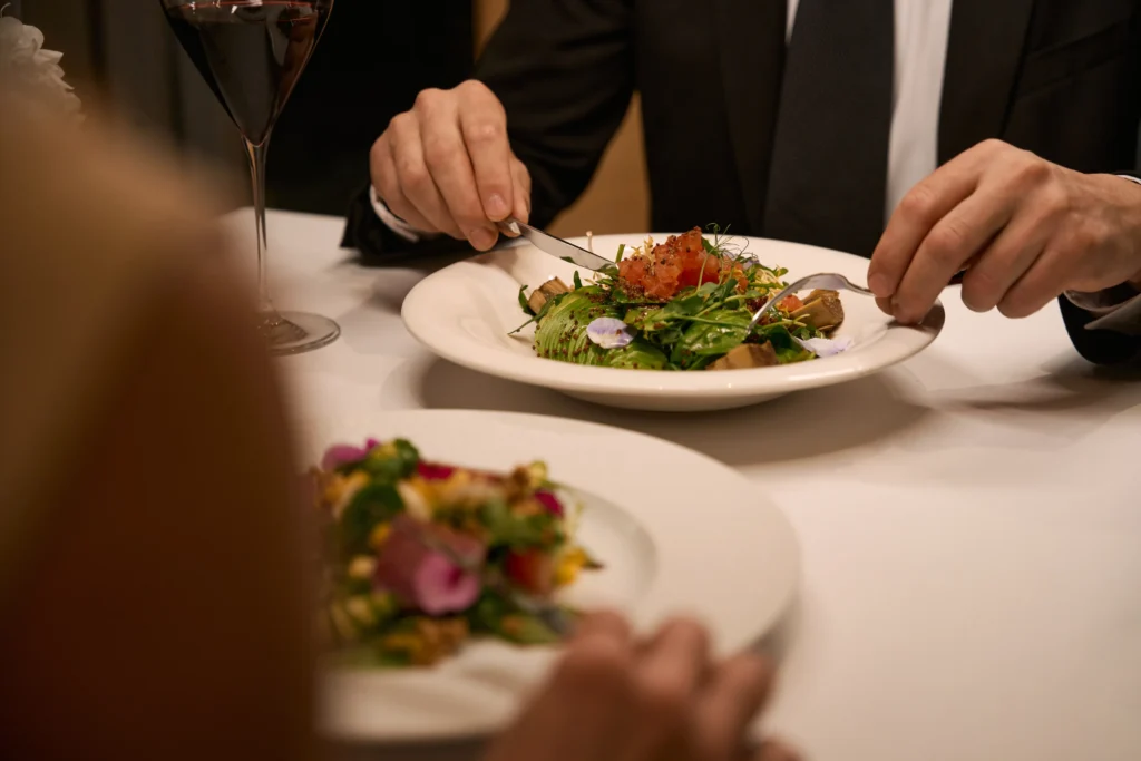 Two people dining at a table, each with a gourmet salad on a white plate. One person in a suit uses a fork and knife. A glass of red wine is visible. The setting appears elegant and formal. Fiamma Sun Valley | Italian