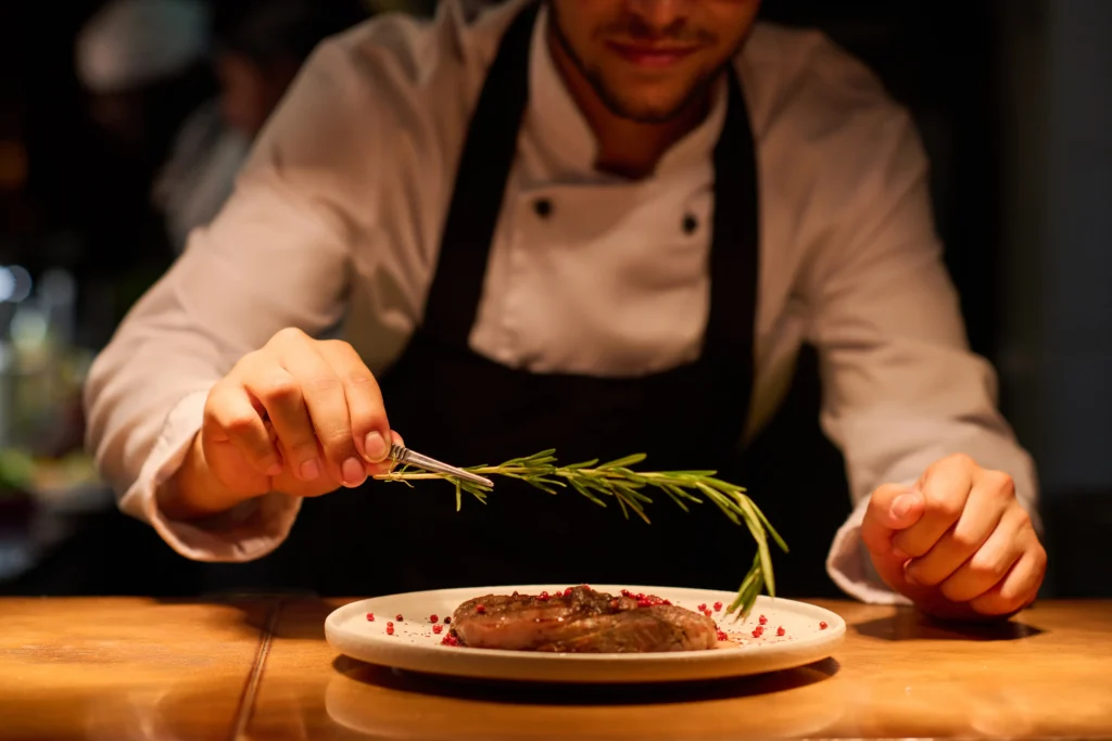 A chef in a white coat garnishes a plated steak with a sprig of rosemary, adding final touches at a wooden counter in a dimly lit kitchen. Fiamma Sun Valley | Italian