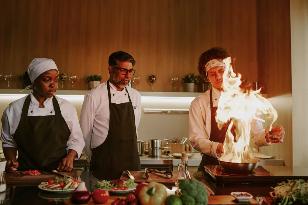 Three chefs in a kitchen watch as one chef creates a large flame while cooking at the stove, with fresh vegetables and ingredients on the counter in front of them. Fiamma Sun Valley | Italian