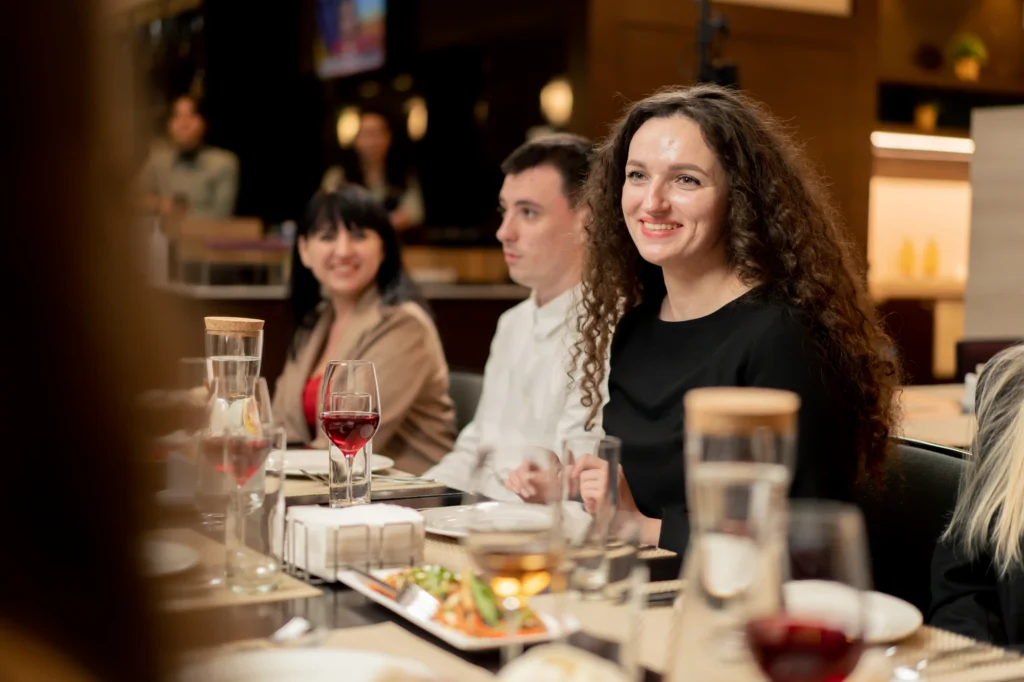 Three people sit at a restaurant table, smiling and talking. There are glasses of wine, water, and plates of food in front of them. The background is softly lit and out of focus. Fiamma Sun Valley | Italian