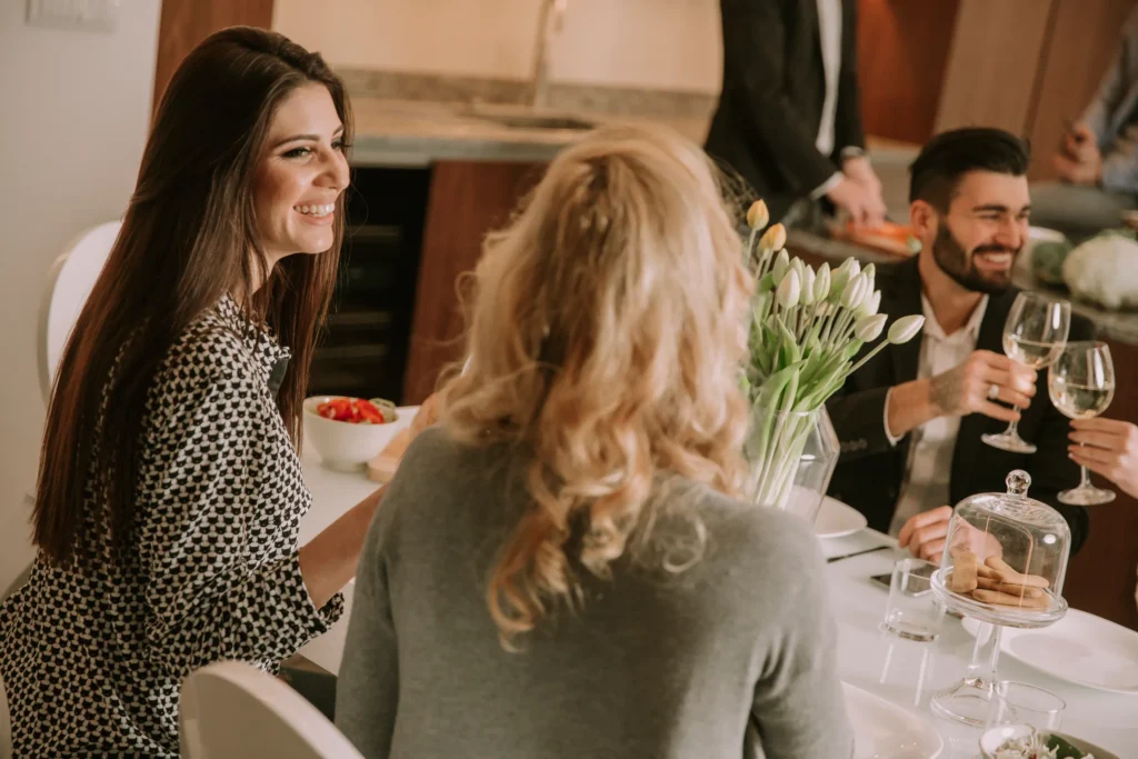 Three people sit at a dining table talking and smiling. One woman holds a bowl of strawberries. There are flowers, wine glasses, and snacks on the table. A man stands in the background near the kitchen. Fiamma Sun Valley | Italian