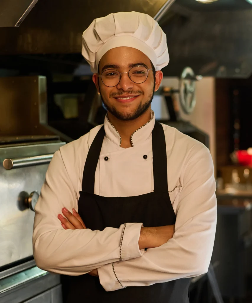 A smiling chef in a white hat, glasses, and uniform stands with arms crossed in a commercial kitchen. Fiamma Sun Valley | Italian