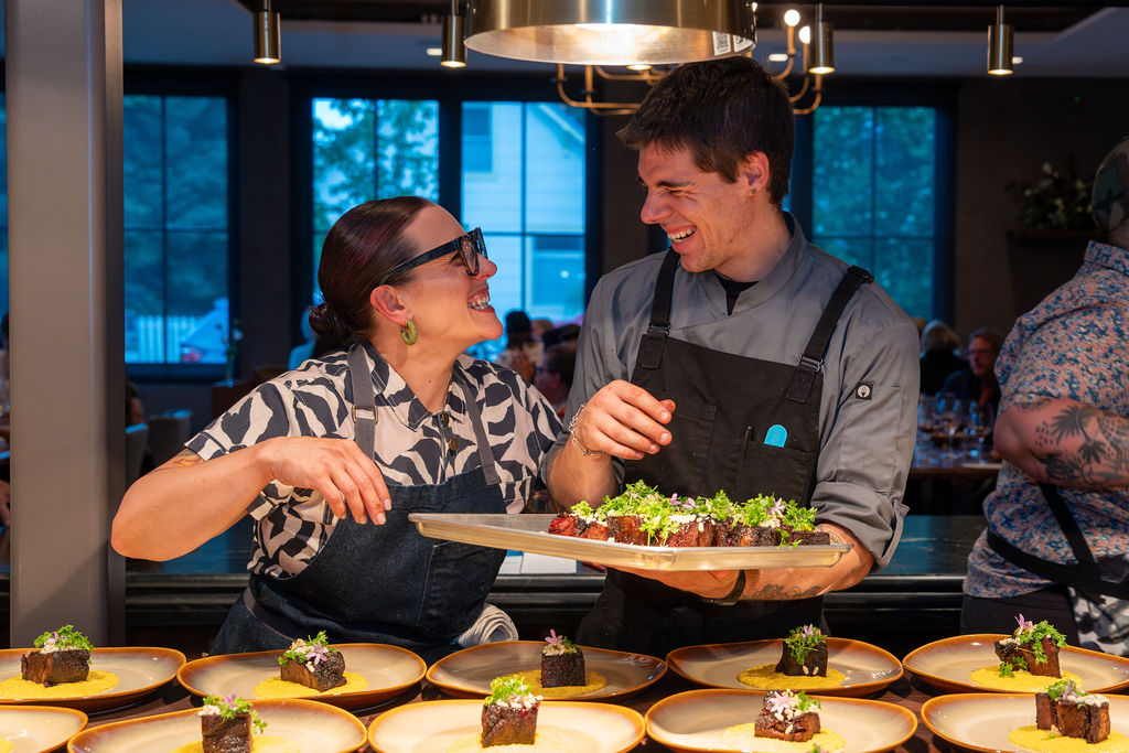 Two smiling chefs stand together, one holding a tray of plated food, surrounded by dishes topped with greens in a bustling restaurant kitchen. Fiamma Sun Valley | Italian