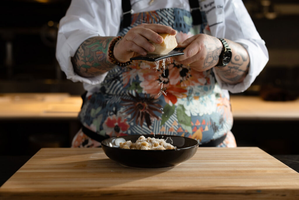 A tattooed chef in a floral apron grates cheese over a bowl of pasta on a wooden board, with cheese shavings falling into the dish. Fiamma Sun Valley | Italian