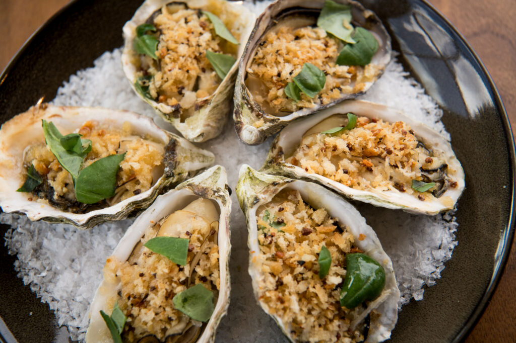 Six baked oysters on the half shell, topped with golden breadcrumbs and fresh green herbs, arranged on a bed of coarse salt in a dark bowl. Fiamma Sun Valley | Italian