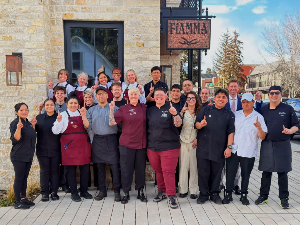 A group of restaurant staff and chefs pose together outside a stone building, smiling and holding up one finger each, with the restaurant sign FAMMA visible above them. Fiamma Sun Valley | Italian