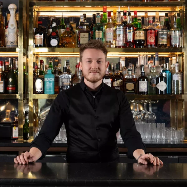 A bartender in a black vest stands behind a bar with shelves of liquor bottles and glassware displayed neatly in the background. Fiamma Sun Valley | Italian