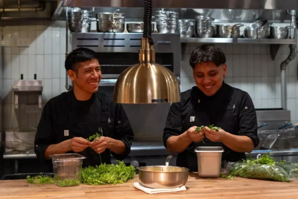 Two chefs in black uniforms smile while preparing fresh herbs at a wooden counter in a professional kitchen with stainless steel appliances and utensils in the background. Fiamma Sun Valley | Italian