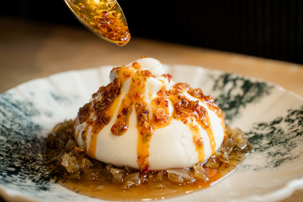 A spoon drizzles chili oil onto a mound of burrata cheese atop a bed of caramelized onions, served on a patterned plate. Fiamma Sun Valley | Italian
