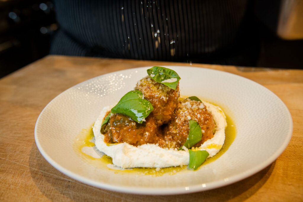 Three meatballs topped with grated cheese and basil sit on a bed of creamy sauce in a white bowl, with olive oil drizzled around, on a wooden table. A person in dark clothing is in the background. Fiamma Sun Valley | Italian