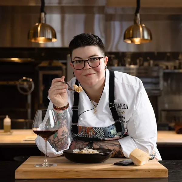 A chef with short dark hair and glasses, wearing a white uniform, sits at a counter holding a forkful of food. A bowl of pasta, a piece of cheese, and a glass of red wine are on the wooden board in front. Fiamma Sun Valley | Italian