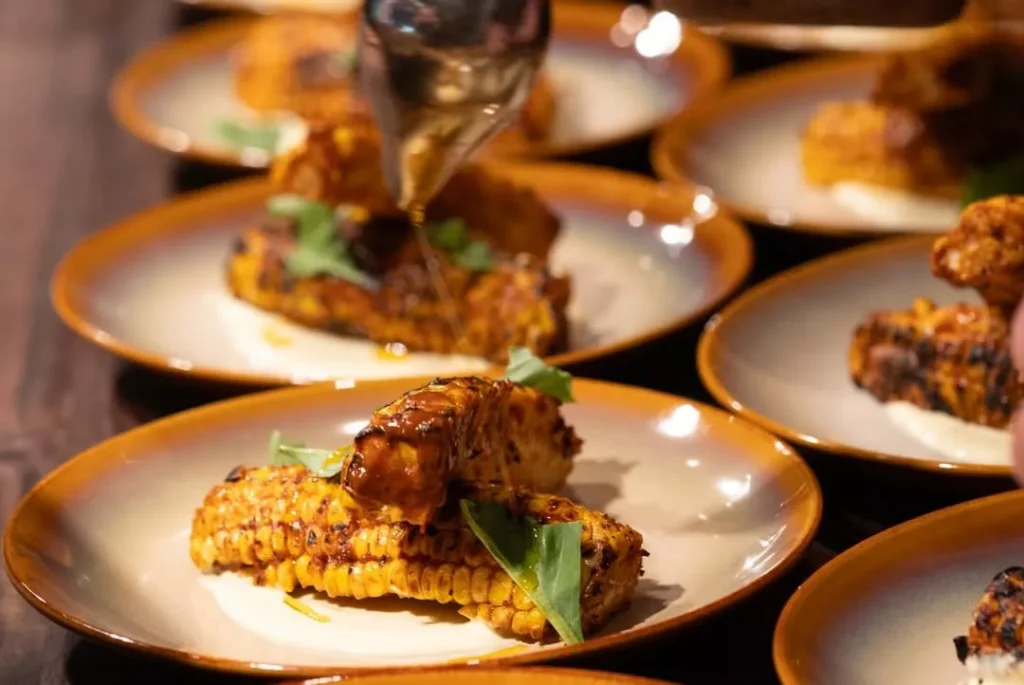 Grilled corn on the cob is served on plates, garnished with greens. A hand is pouring sauce over the corn. The corn is charred and arranged neatly, with several plates visible on a wooden table. Fiamma Sun Valley | Italian