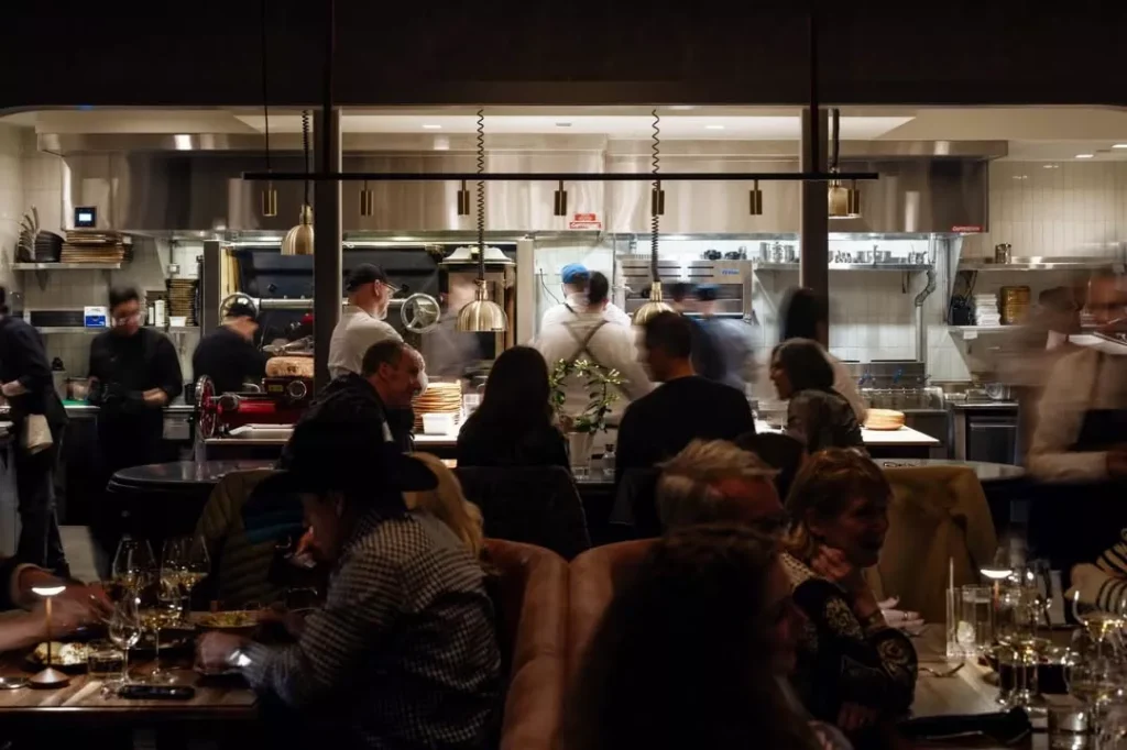 A bustling restaurant scene shows diners seated at tables in the foreground, while chefs and staff work in an open kitchen in the background under warm lighting. Fiamma Sun Valley | Italian