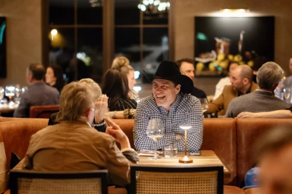 A man in a black cowboy hat laughs while dining with others at a warmly lit restaurant. Multiple groups are seated at tables, engaged in conversation. Wine glasses and table lamps are visible. Fiamma Sun Valley | Italian