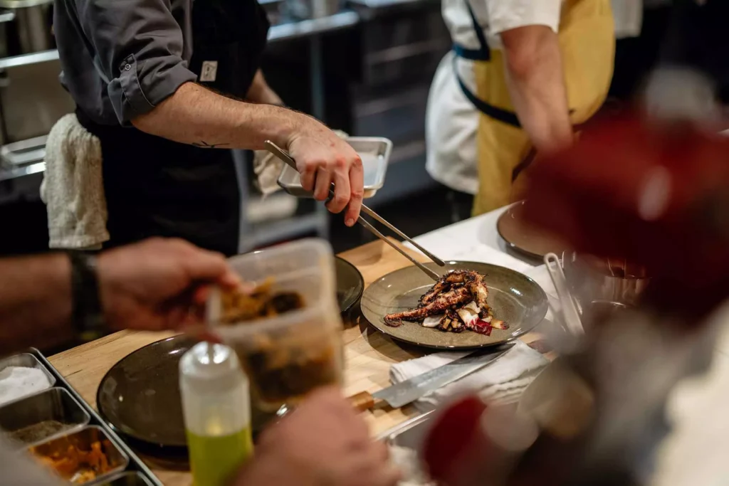 A chef uses tongs to plate grilled octopus in a busy kitchen, surrounded by other chefs, food containers, and kitchen tools on a wooden counter. Fiamma Sun Valley | Italian