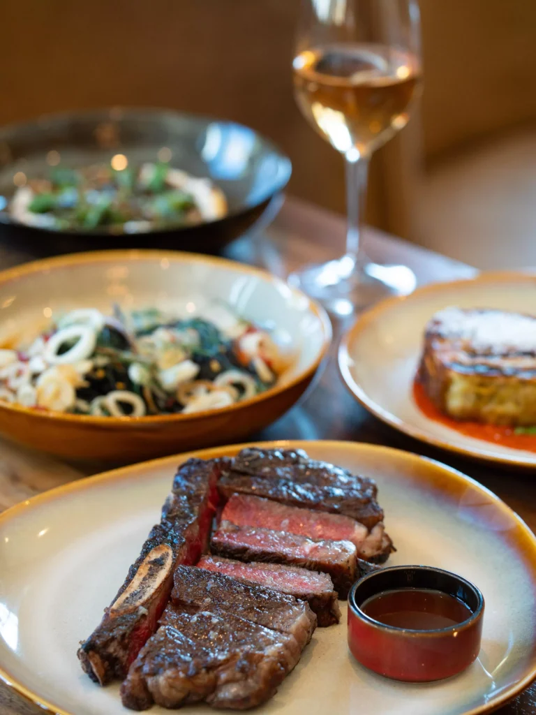 A plate with sliced steak and sauce, a bowl of pasta, another dish with greens, a dessert, and a glass of white wine on a wooden table. Fiamma Sun Valley | Italian