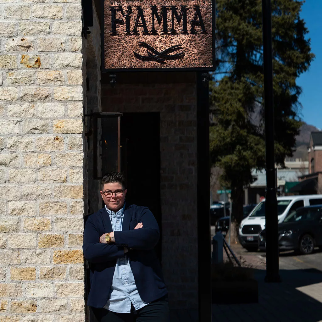 A person in glasses and a dark jacket leans against a light brick wall under a sign that says FIAMMA on a sunny street with parked cars and tall trees in the background. Fiamma Sun Valley | Italian