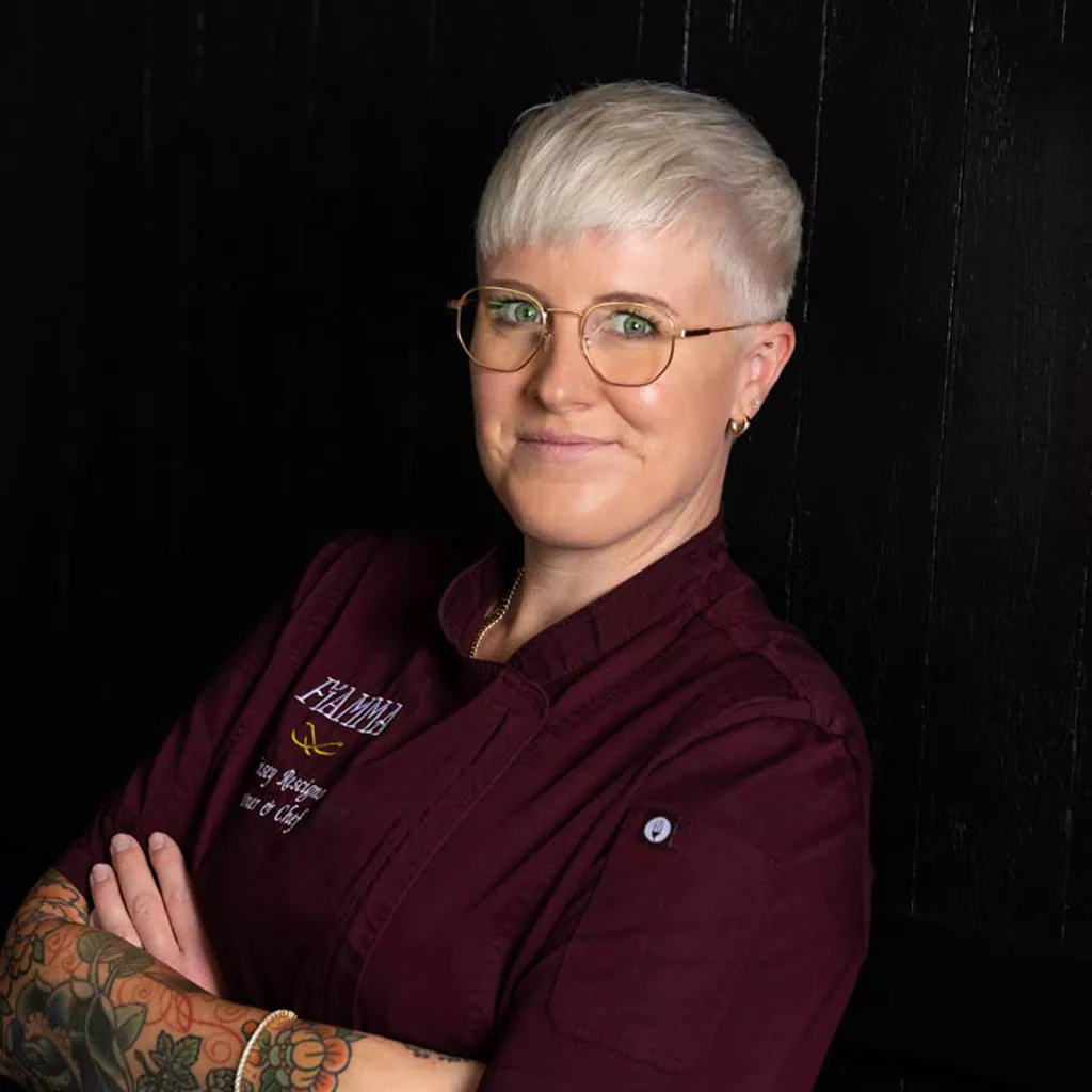 Person with short platinum blonde hair, glasses, and tattoos, wearing a burgundy chefs jacket, stands with arms crossed and smiles slightly in front of a dark background. Fiamma Sun Valley | Italian