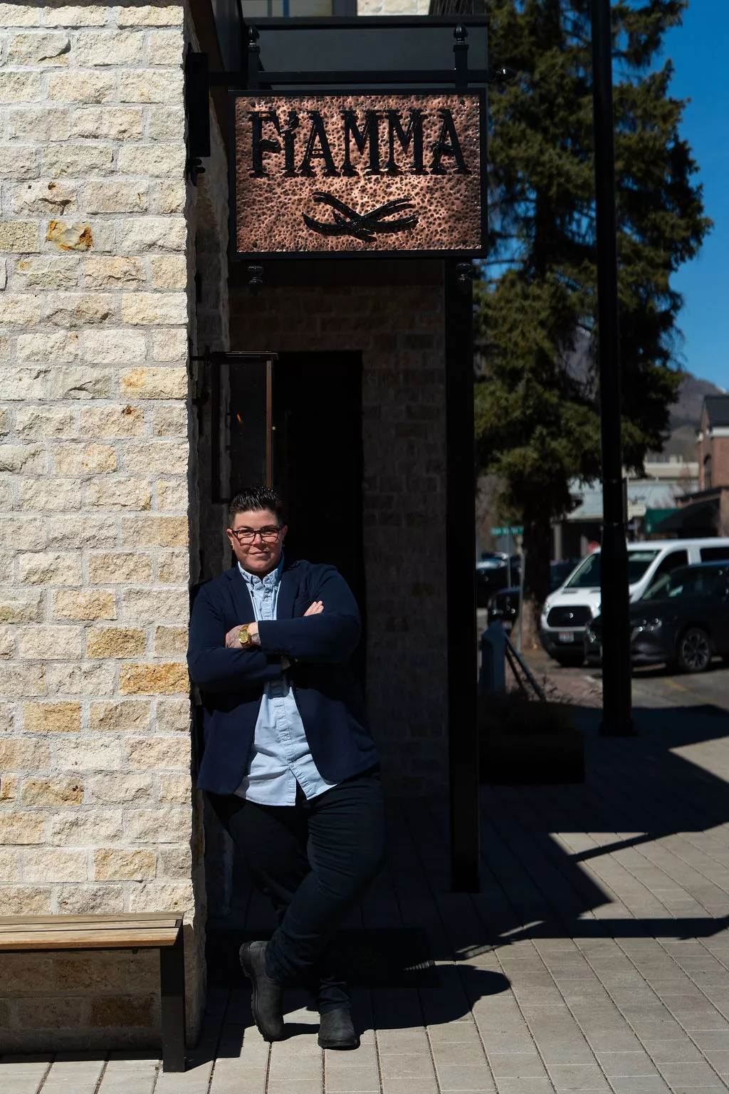 A person with short hair, glasses, and a blazer leans with arms crossed against a stone wall beneath a Fiamma sign on a sunny sidewalk. Britt Rescigno stands confidently, while cars and trees are visible in the background. Fiamma Sun Valley | Italian