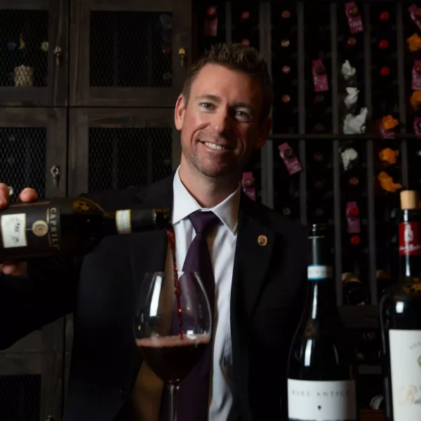 A smiling man in a suit pours red wine into a glass at a wooden table, with wine bottles and shelves of wine in the background, creating an inviting scene reminiscent of Britt Rescigno’s sophisticated gatherings. Fiamma Sun Valley | Italian