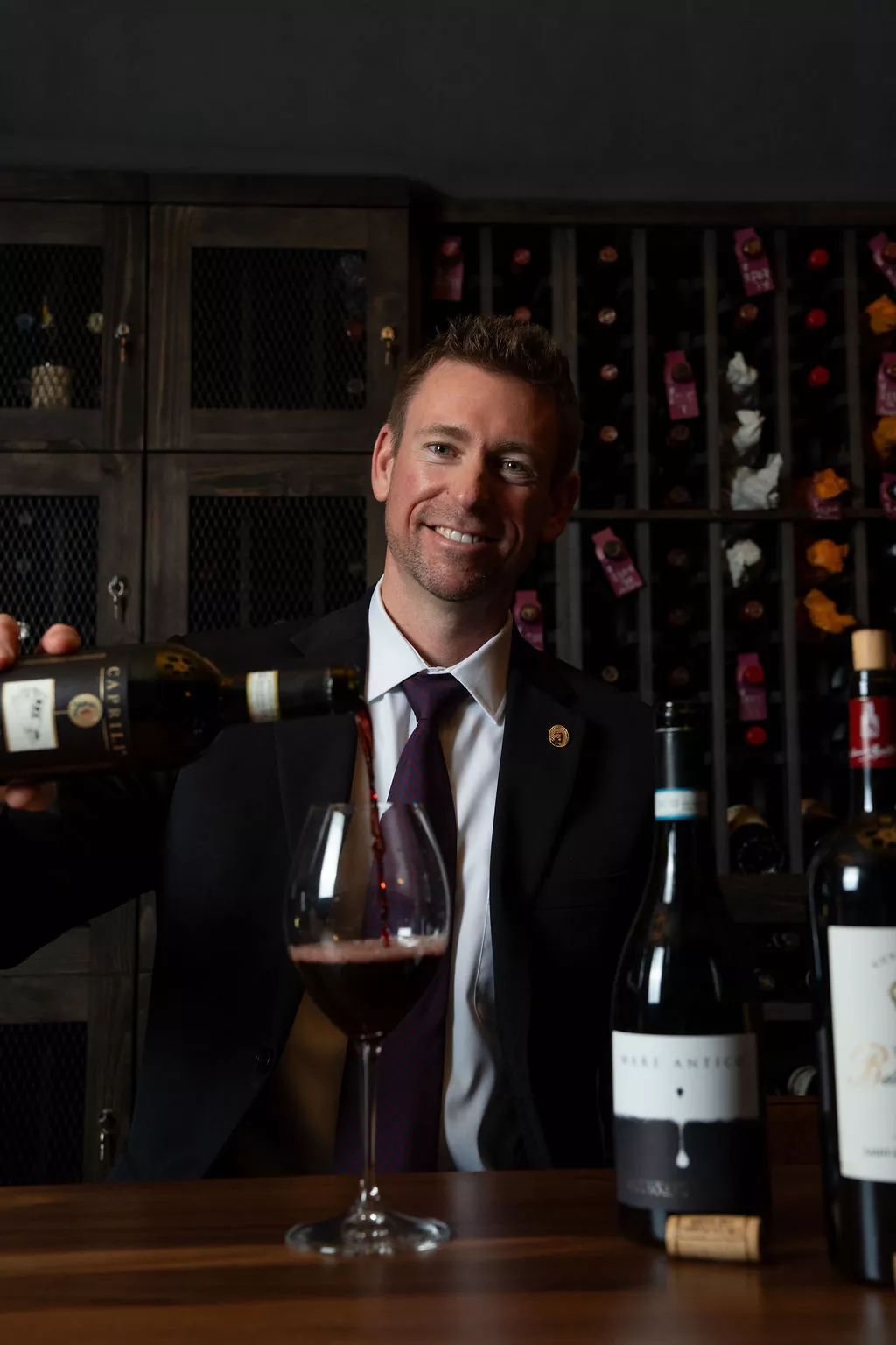 A smiling man in a suit pours red wine into a glass at a wooden table, with wine bottles and shelves of wine in the background, creating an inviting scene reminiscent of Britt Rescigno’s sophisticated gatherings. Fiamma Sun Valley | Italian