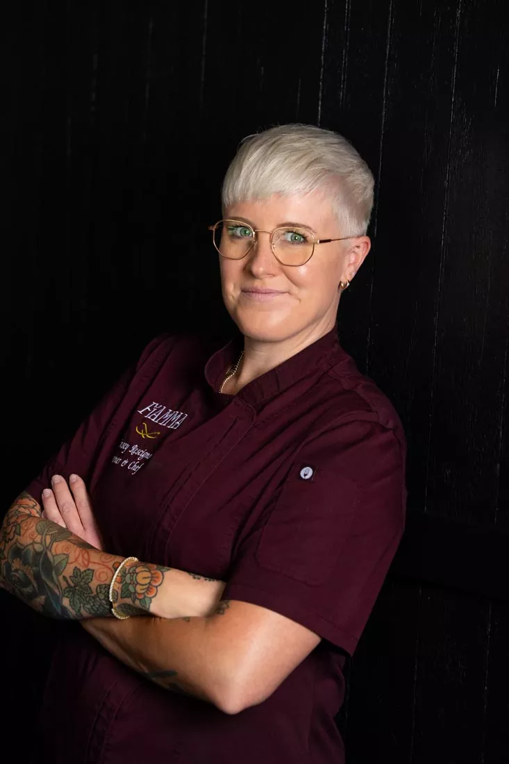 A person with short white hair, glasses, and tattoos on their arms stands with folded arms, wearing a dark burgundy shirt, in front of a black wooden background, smiling slightly—a look reminiscent of Britt Rescigno’s confident and approachable style. Fiamma Sun Valley | Italian