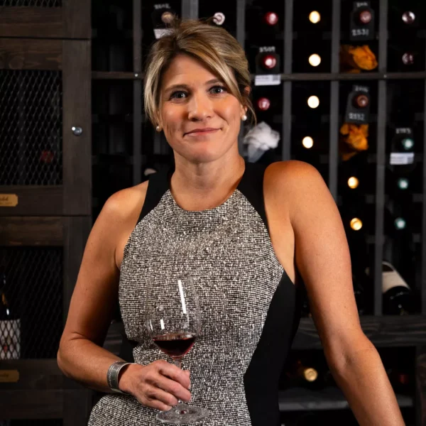 A woman in a sleeveless dress holds a glass of wine and smiles, standing in front of shelves filled with wine bottles. Fiamma Sun Valley | Italian