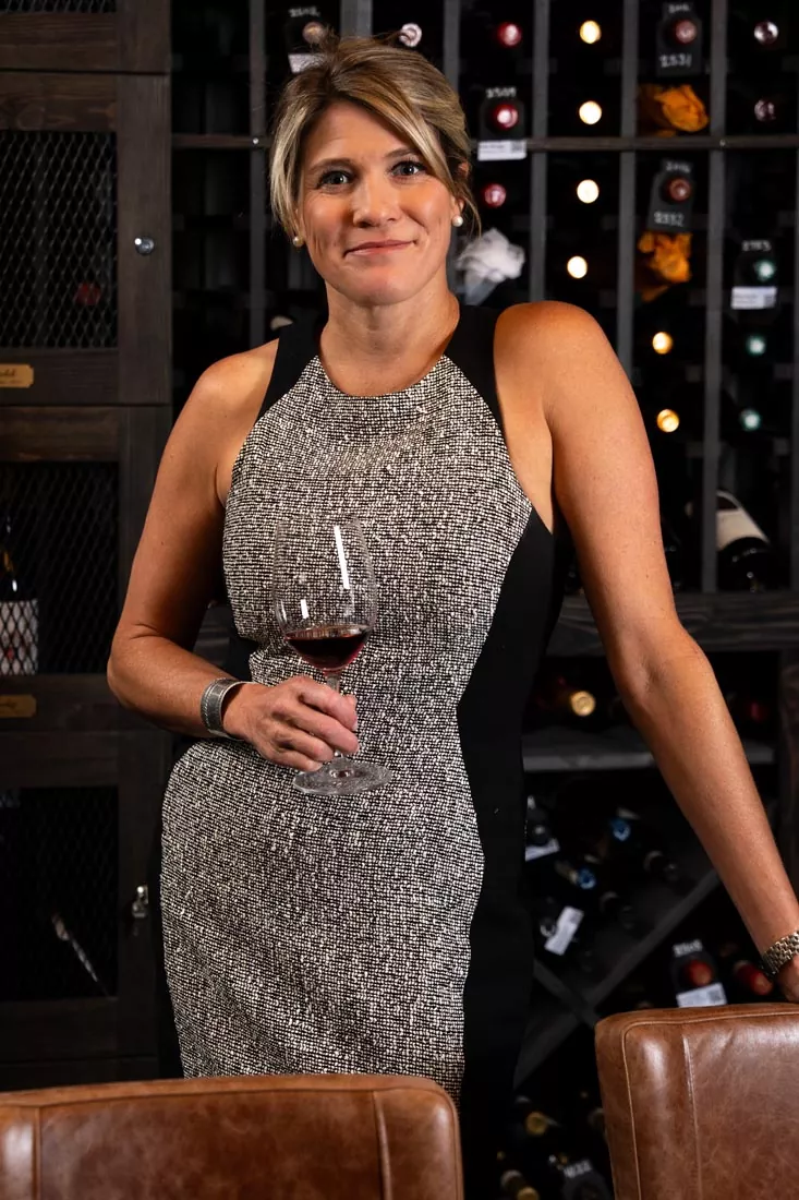 A woman in a sleeveless gray and black dress holds a glass of red wine and smiles, standing in front of a wine rack filled with bottles—capturing the elegant, confident style often associated with Britt Rescigno. Fiamma Sun Valley | Italian