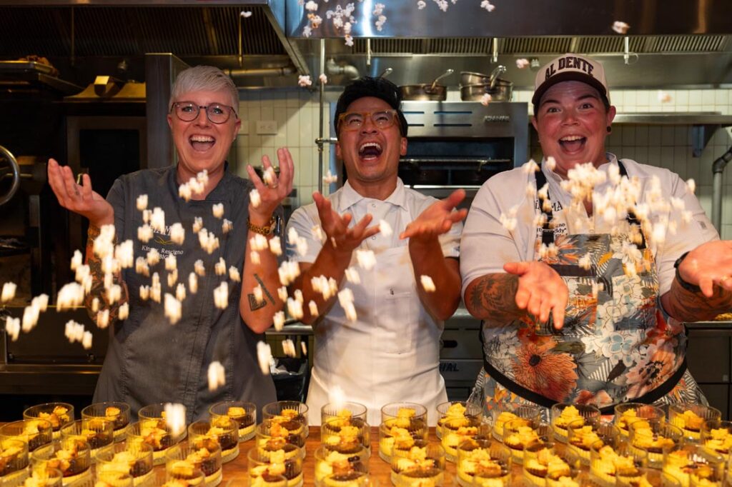 Three chefs in a kitchen joyfully toss popcorn in the air over a table filled with dessert cups, smiling and celebrating together—among them is Britt Rescigno, adding her signature flair to the festive scene. Fiamma Sun Valley | Italian